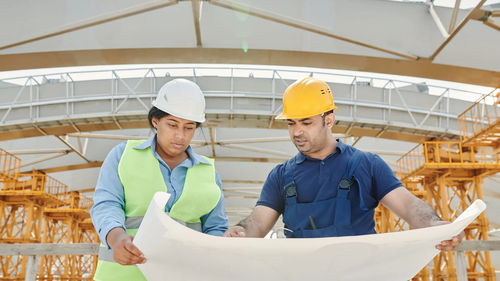 Two engineers in hard hats reviewing a large printed plan together under a steel roof structure